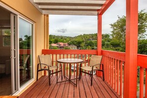 Een houten terras met een glazen deur naar een kamer, een ronde tafel met stoelen en een rode balustrade.
