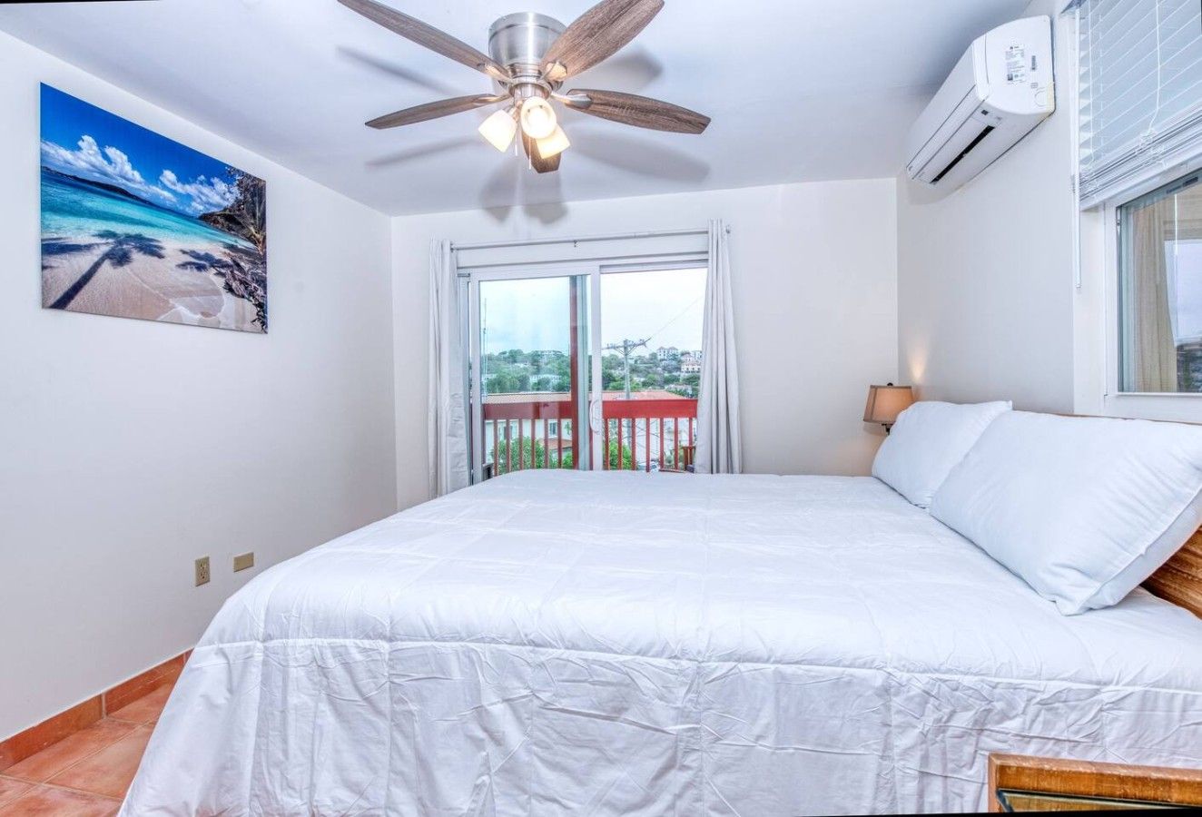 Un dormitorio con cama, ventilador de techo, equipo de aire acondicionado, ventana con cortinas y un cuadro con una escena de playa.