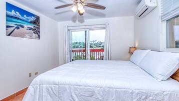 Un dormitorio con cama, ventilador de techo, equipo de aire acondicionado, ventana con cortinas y un cuadro con una escena de playa.