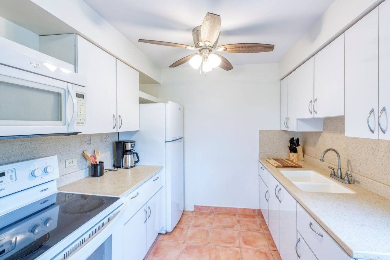 Cocina compacta con gabinetes blancos, microondas, estufa, cafetera y ventilador de techo.