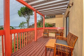 A wooden deck with a red railing, a table, and two chairs, overlooking a coastal area with vegetation and a building in the background.
