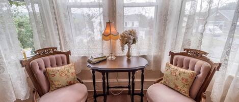 A room with two wooden chairs with floral cushions, a small table with a lamp, and lace curtains.