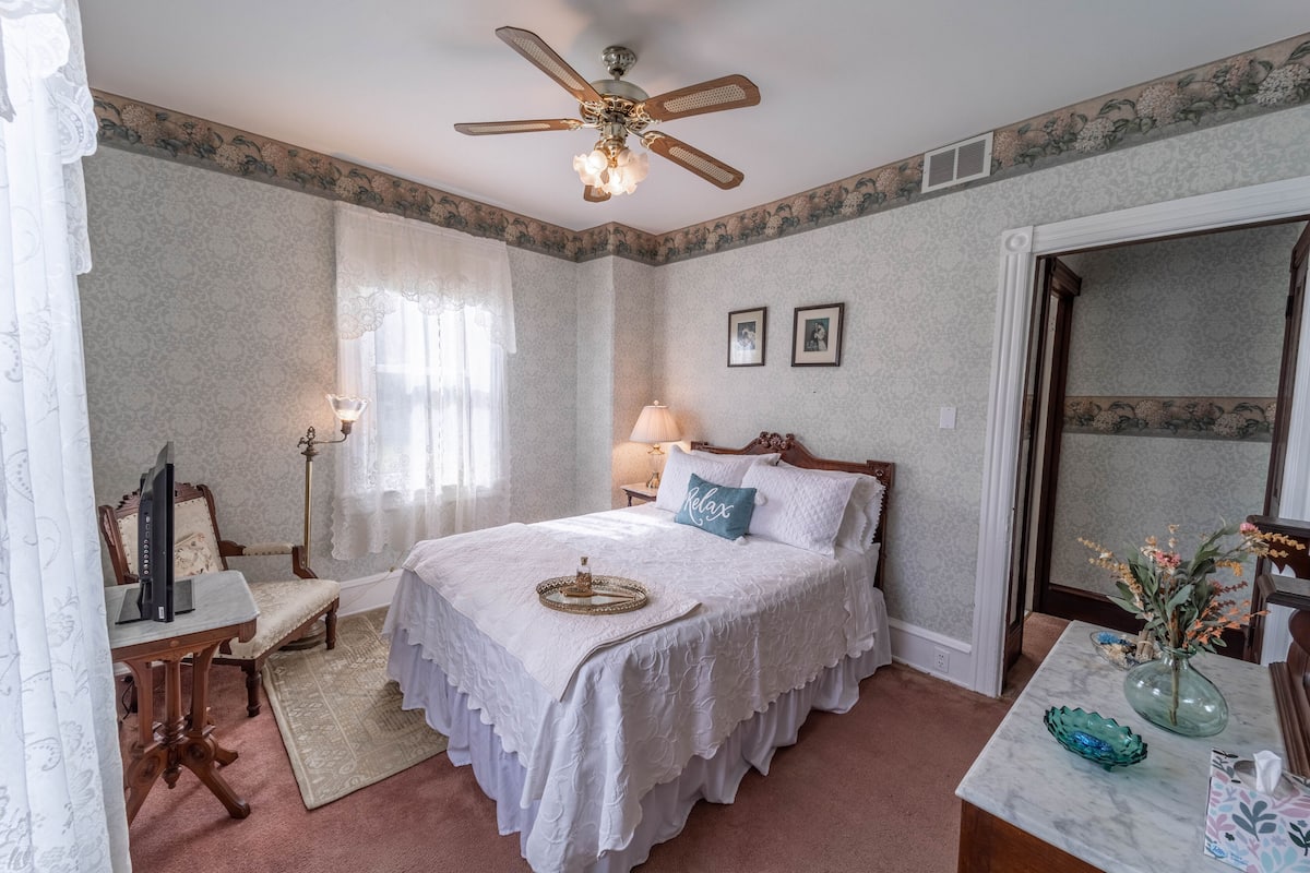 Un dormitorio con cama, escritorio, silla, lámpara de pie, ventilador de techo y un florero con flores.