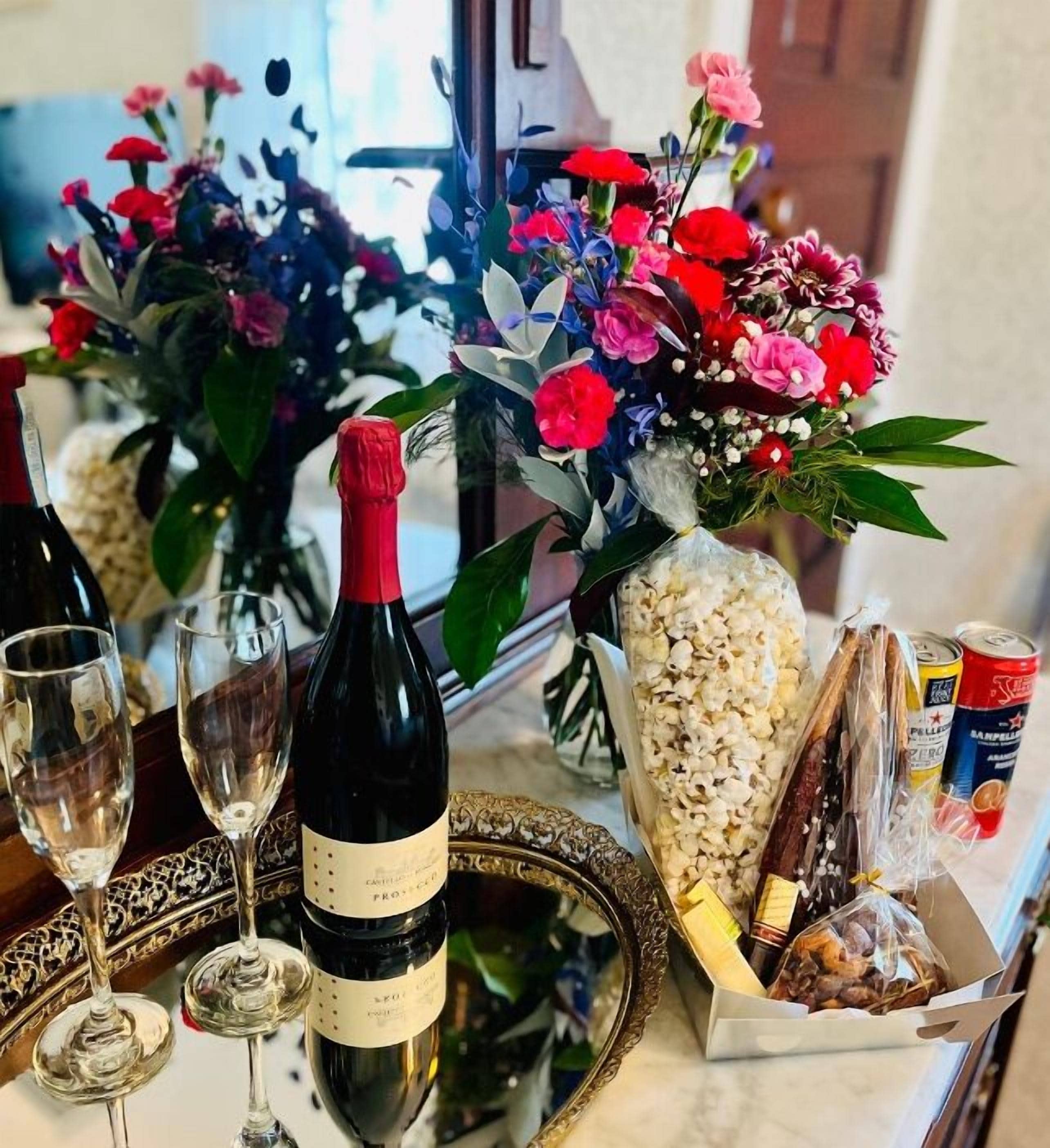 A table with a bottle of wine, two wine glasses, a bouquet of flowers, a basket of snacks, and a can of soda.