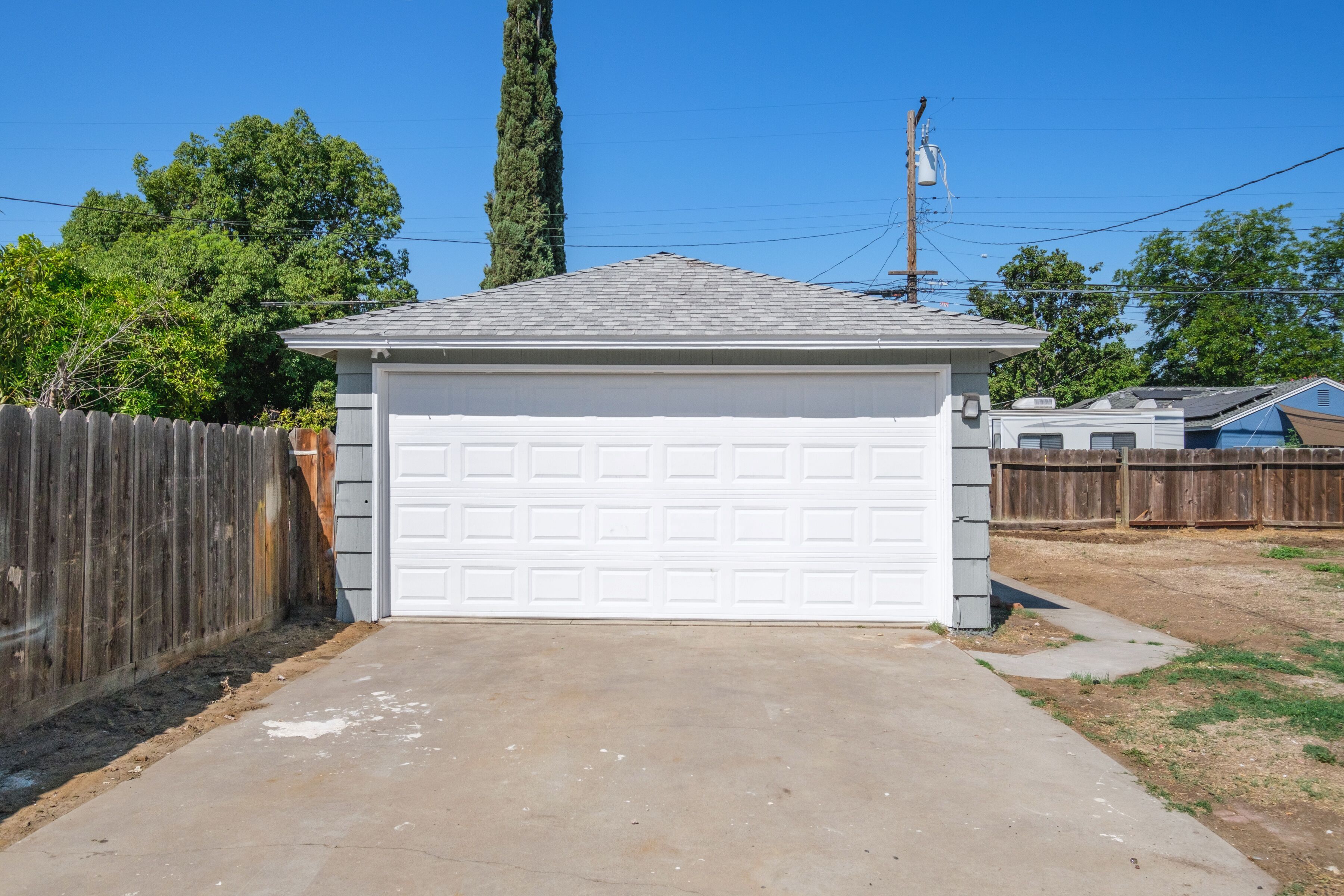 Un garage avec une porte blanche, une allée en béton, une clôture en bois et un grand arbre en arrière-plan.