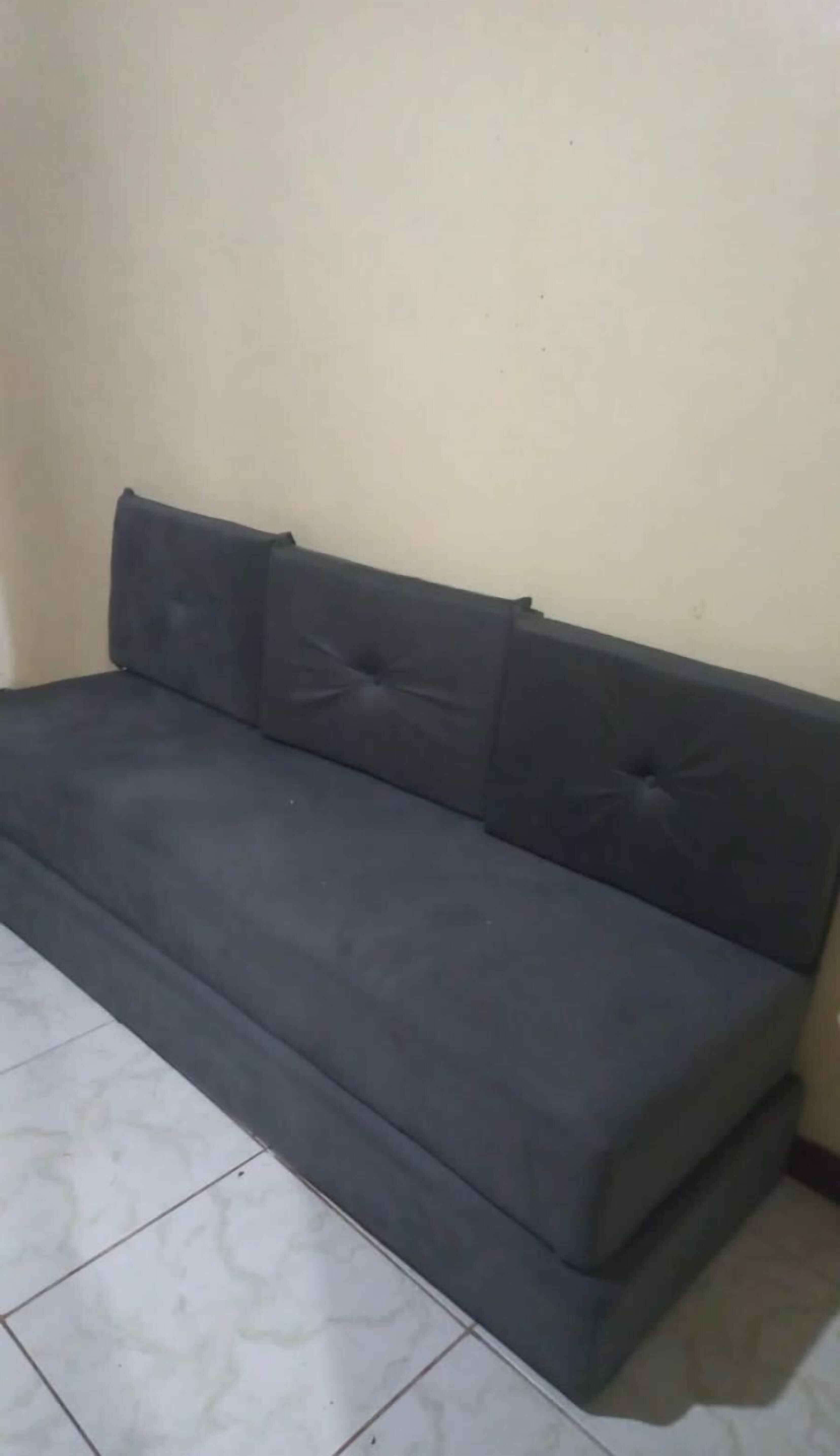 A dark-colored sofa with a tufted backrest in a room with plain walls and tiled flooring.