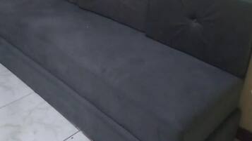 A dark-colored sofa with a tufted backrest in a room with plain walls and tiled flooring.