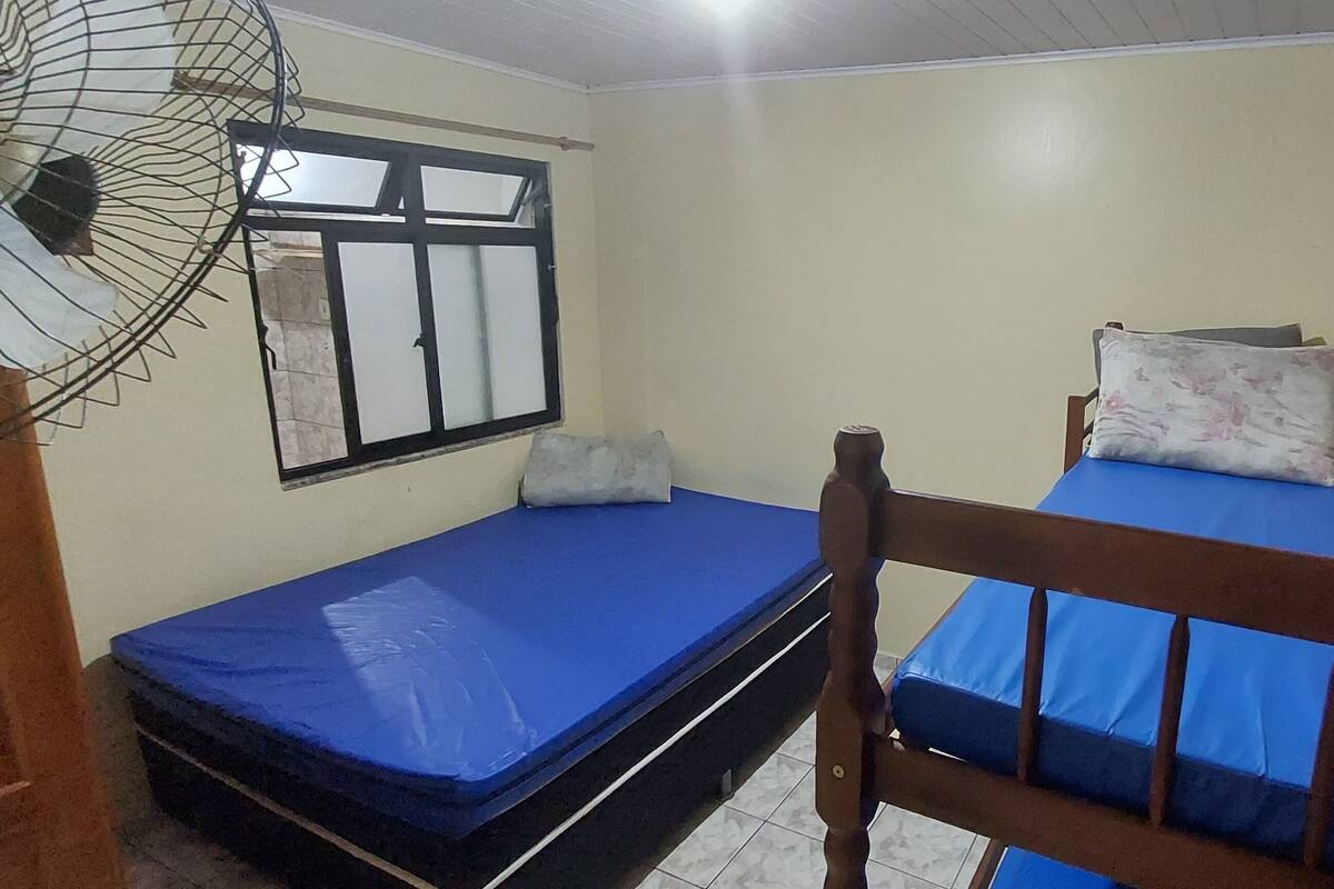 Ein kleines Zimmer mit zwei Einzelbetten, einem Fenster mit Metallrahmen und einem Standventilator.