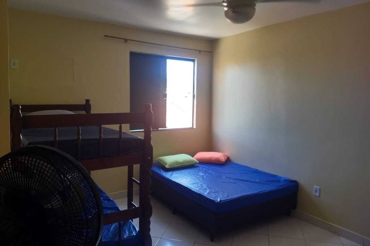Ein Zimmer mit Etagenbett, einem Einzelbett, einem Fenster mit Tageslicht, einem Deckenventilator und einem Standventilator.