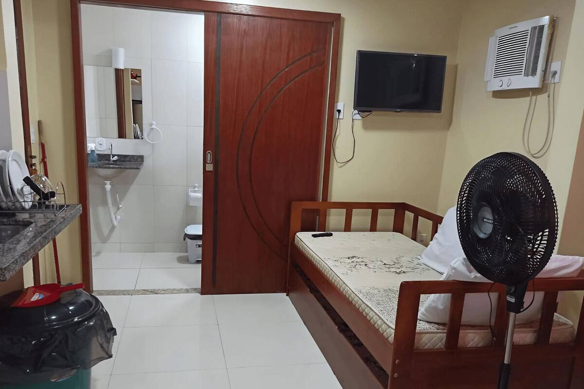 Ein kleines Zimmer mit Bett, Ventilator, Fernseher und einem durchgegangenen Tür angedeuteten Badezimmer.