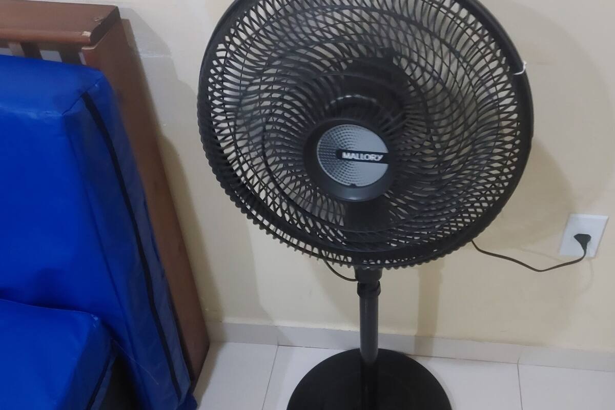 Ein Standventilator mit runder Schutzgitterabdeckung und Standfuß, platziert vor einer Wand mit einer Steckdose in der Nähe.