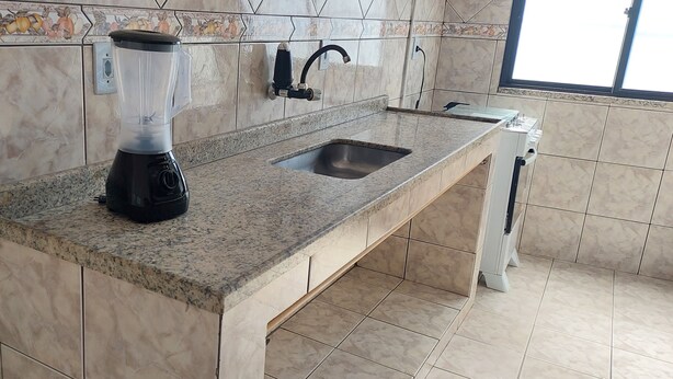 A kitchen with a granite countertop, a built-in sink, a blender, and a tiled backsplash with a fruit pattern. - Brisas do Farol - Aluguel Economico (Arraial do Cabo)
