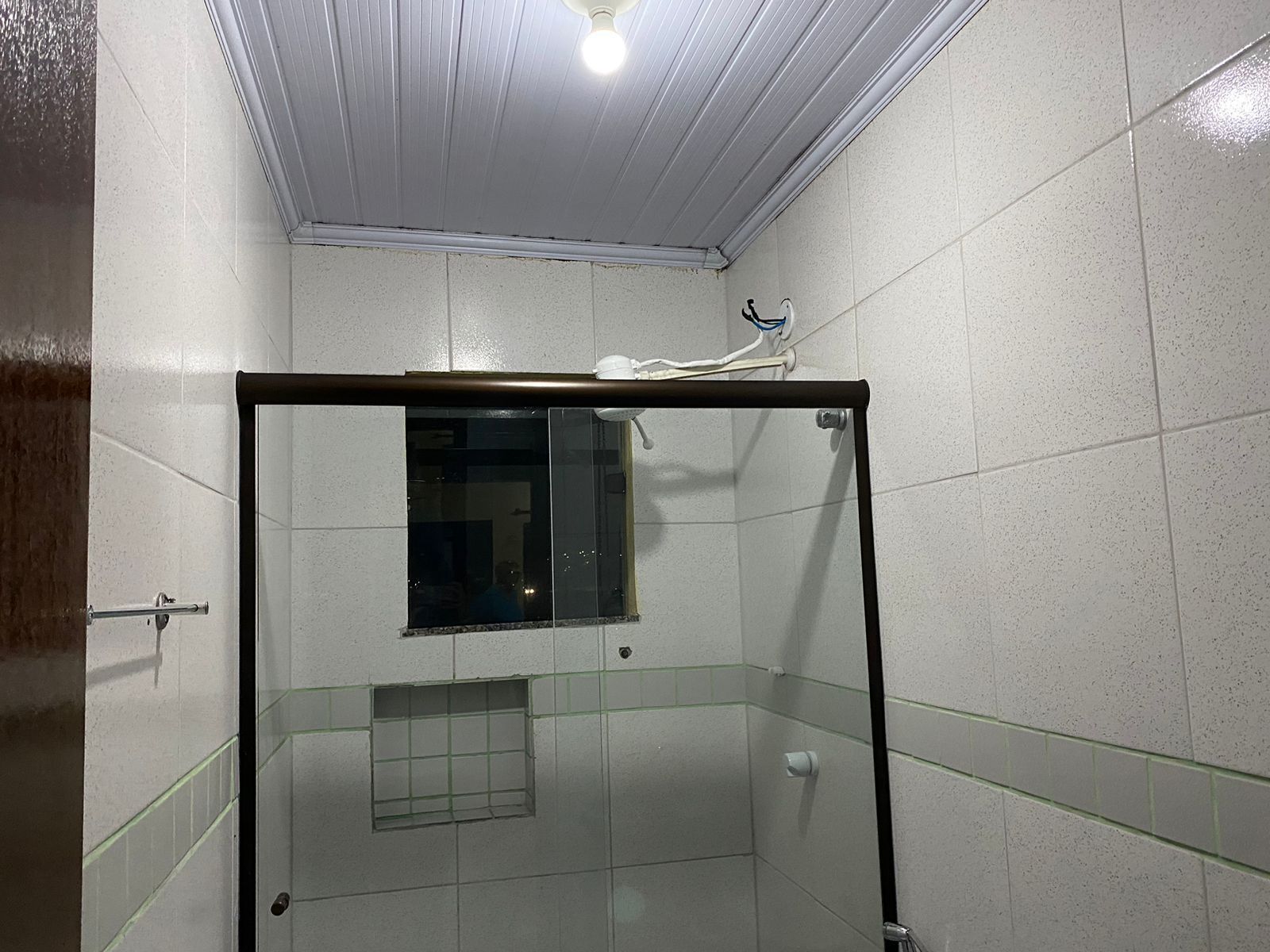 Una ducha con cerramiento de vidrio, estructura metálica, paredes de azulejos blancos y una lámpara visible en el techo.