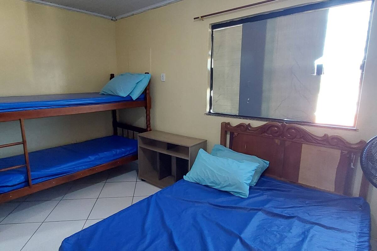 Ein Zimmer mit zwei Etagenbetten, einem Einzelbett, einem Standventilator, einem Fenster und einem Spiegel.