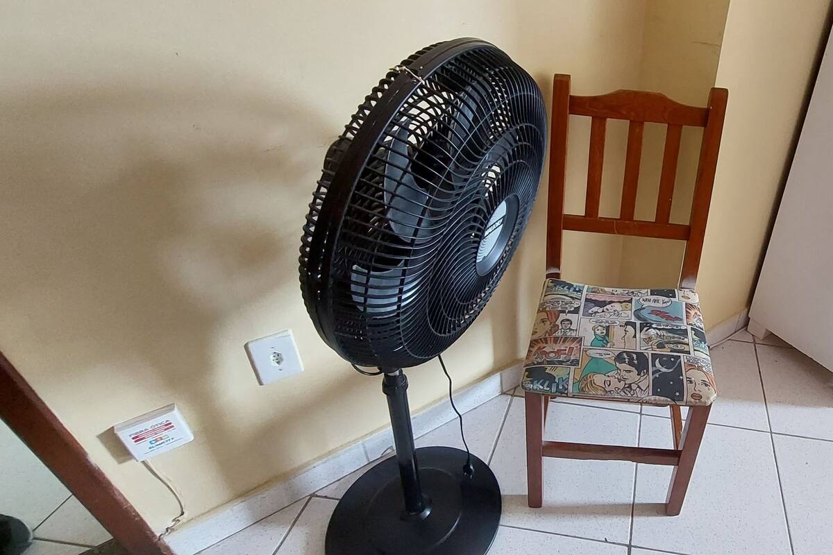 Ein Zimmer mit einem Standventilator, einem Holzstuhl mit farbigem Kissen und einem gefliesten Boden.