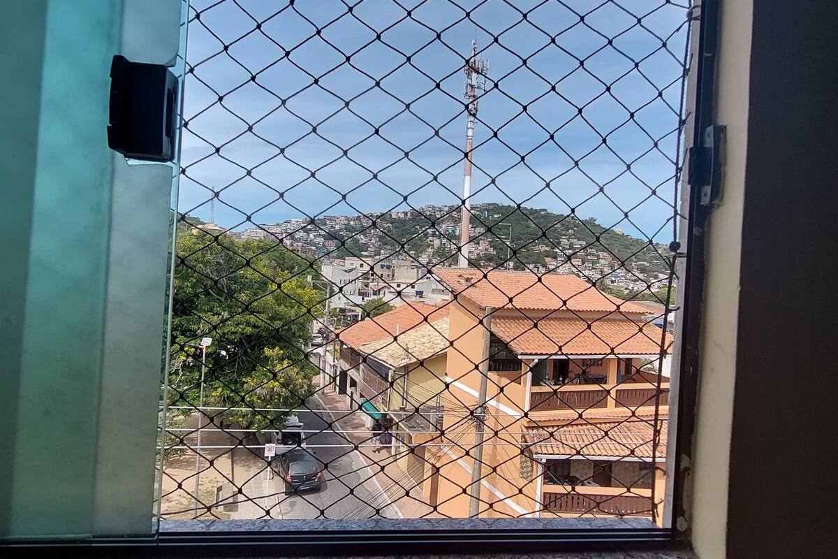 Blick durch ein Fenster mit einem Maschendrahtzaun, der eine Straße mit Gebäuden, einen Hügel und einen hohen Sendeturm zeigt.