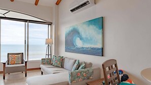 A room with a large window, a painting of a wave, a sofa, a chair, and a small table. - Sunset 303 - Ocean View 3 BR Condo (Potrero)