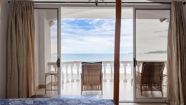 A bedroom with a large bed, a sliding glass door leading to a balcony with wicker chairs and a view of the sea. - Sunset 303 - Ocean View 3 BR Condo (Potrero)
