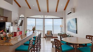 A modern kitchen with a large island, a dining area with a round table and chairs, and a large window offering a view of the sea. - Sunset 303 - Ocean View 3 BR Condo (Potrero)