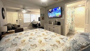 Un dormitorio con cama, ventilador de techo, televisión, cómoda y baño con cortina de ducha.