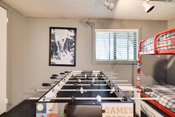 Una habitación con mesa de futbolín, literas, ventana con persianas y un cuadro en la pared.