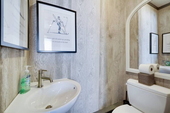A bathroom with a white sink, a framed illustration of a skier, and a toilet with a roll of toilet paper.