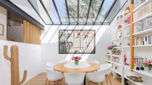 A modern dining area with a round wooden table surrounded by white chairs, a cactus decoration, and a bookshelf with various items.