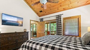Un dormitorio con una colcha a cuadros, techo de madera, ventilador de techo, cómoda, un cuadro en la pared y vistas al exterior a través de una puerta.