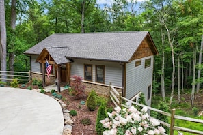 Situated in a natural forest setting, this cabin features a front porch and a garden with various shrubs and flowering plants.