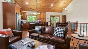 Amplia sala de estar con sofá de cuero y una mesa de centro, cocina con armarios de madera y comedor con vistas al exterior.