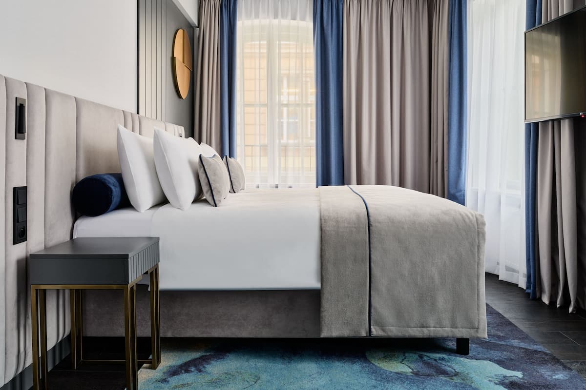 Una habitación de hotel moderna con una cama grande, una mesita de noche, un televisor de pantalla plana y vistas de la ciudad a través de amplios ventanales.