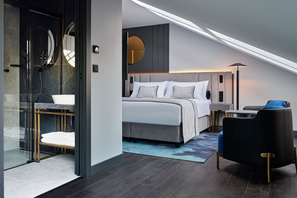 Una habitación de hotel moderna con cama, zona de estar, ducha con cerramiento de vidrio y un espejo redondo.