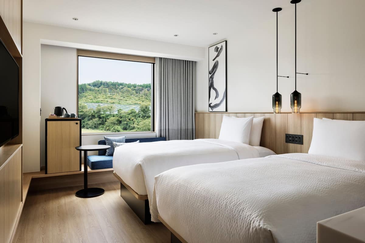 A hotel room with two beds, a TV, a small table, and a window with a view of greenery.