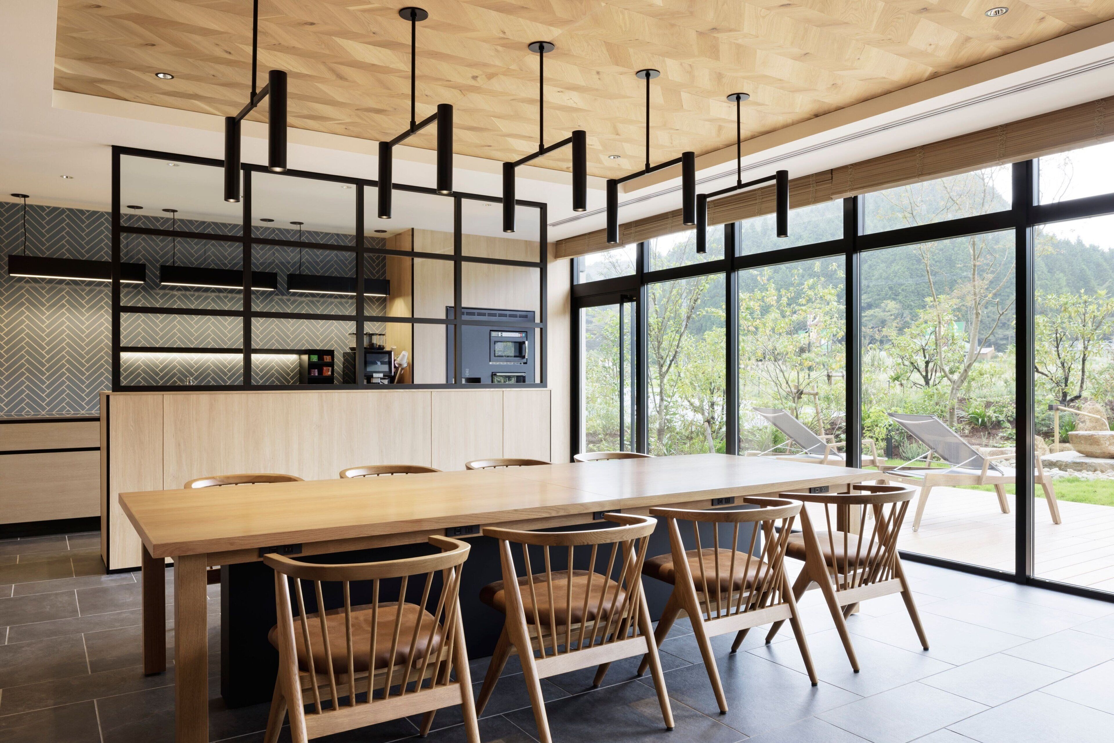 A modern dining area with a long wooden table and chairs, a kitchen with a built-in oven and microwave, and large windows offering a view of greenery.