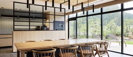 A modern dining area with a long wooden table and chairs, a kitchen with a built-in oven and microwave, and large windows offering a view of greenery.