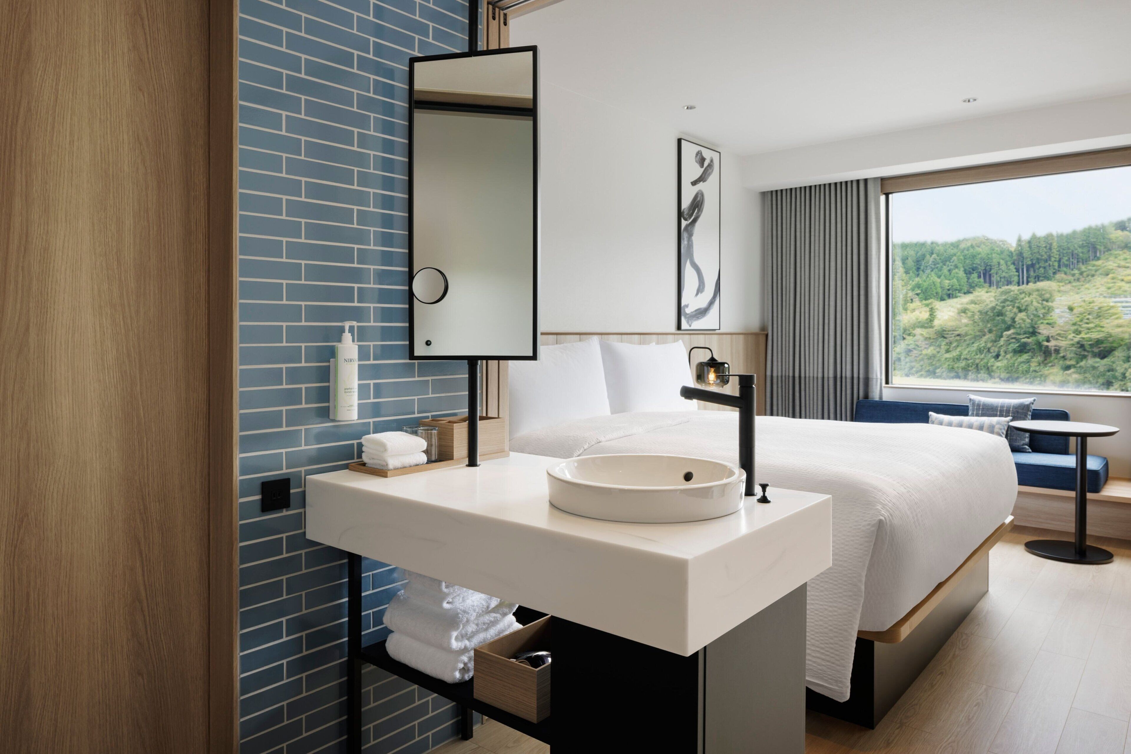A hotel room with a large bed, a white sink, a mirror, and a window with a view of trees.