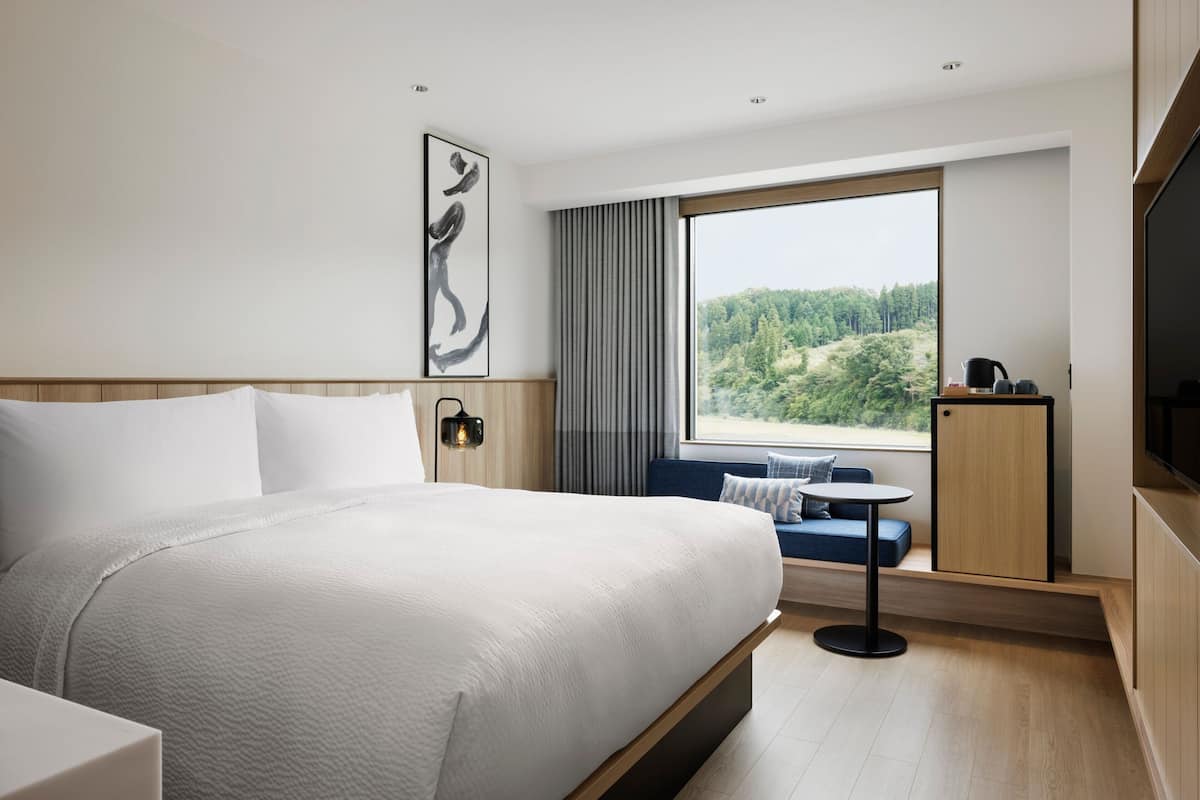 A modern hotel room with a large bed, a small table, a sofa, a TV, and a window with a view of a forest.