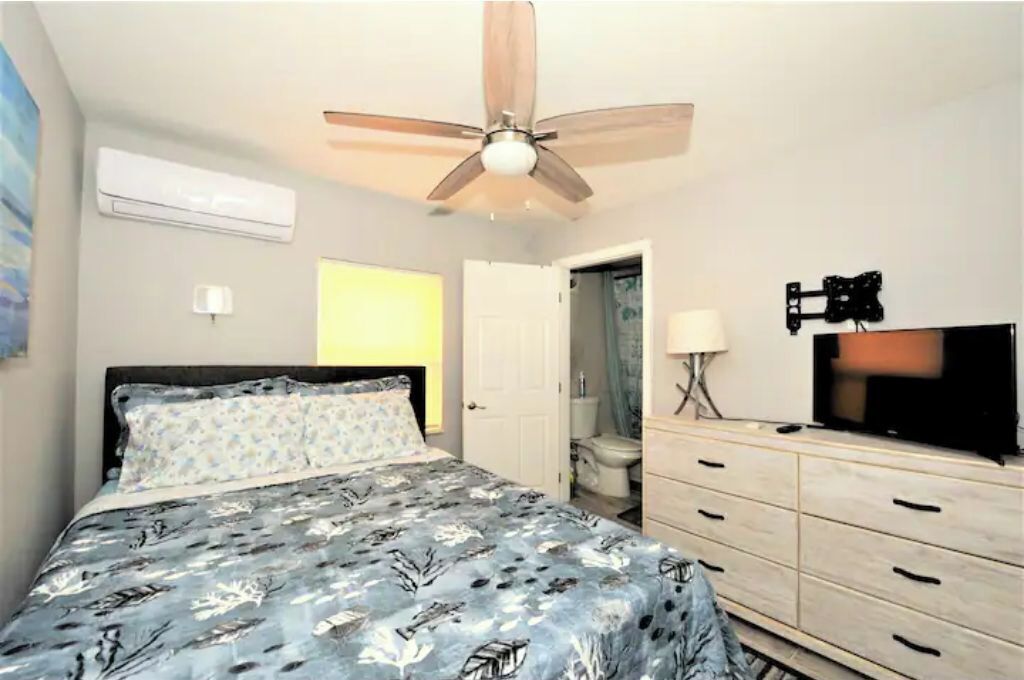 A bedroom with a bed, a ceiling fan, a TV, a dresser, and a bathroom door.