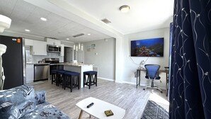 Ein modernes Wohnzimmer mit einer Couch, einem Couchtisch und einem Fernseher. Es gibt eine Küchenzeile mit einer Mikrowelle und einem Kühlschrank.