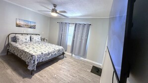 Ein Schlafzimmer mit Bett, Deckenventilator, einem Fenster mit Vorhängen und einem an der Wand montierten Fernseher.