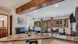 A kitchen with a granite countertop, a wine bottle, and a bar stool. There is a dining area with a couch and a TV mounted on a stone wall. - 🏔️Ski Access~Hot Tub~Studio Premier 102🏔️ (Snowmass Village)