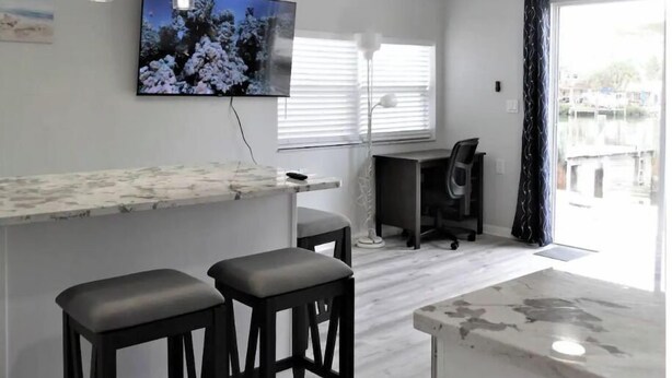 Una cocina moderna con encimera de mármol, dos taburetes de bar, un televisor en la pared y una ventana con persianas.