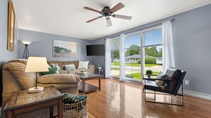 Ein Wohnzimmer mit Deckenventilator, einer Couch, einem Couchtisch und einem Sessel. Ein Fenster bietet Blick auf ein Haus und Grünflächen.