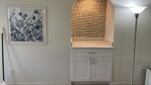 Een kamer met een wit kastje, een lijst met bloemkunst, een metselwerk achterwand en een vloerlamp.