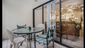 Een glazen patio met een ronde tafel en stoelen, een plafondventilator en uitzicht op een woonkamer met een bank en sierplanten.