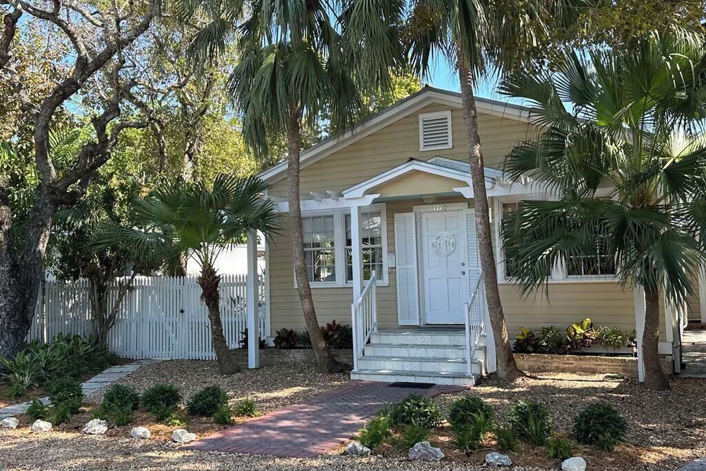 A single-story house with a front porch, a white picket fence, and palm trees.