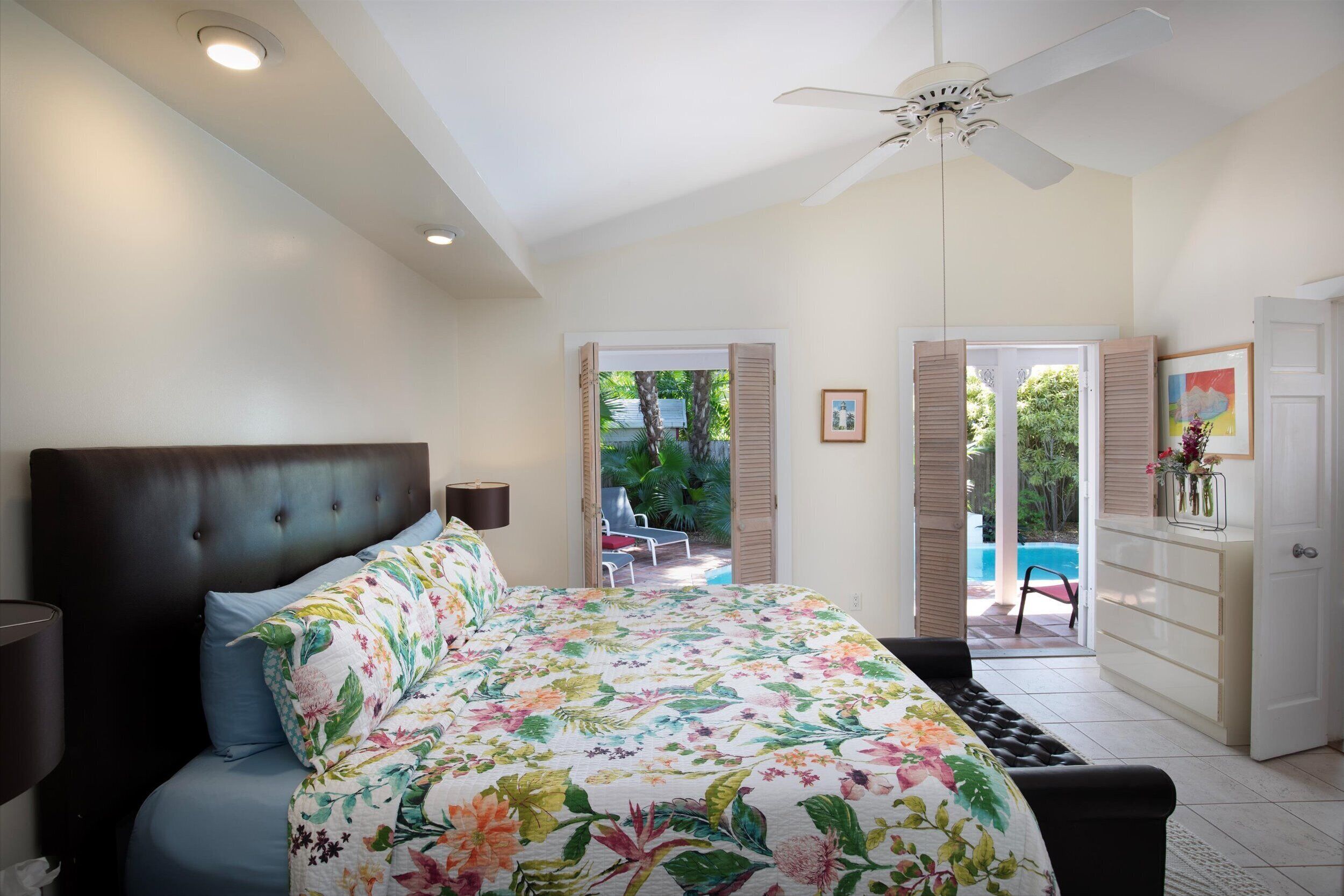 A bedroom with a large bed, a ceiling fan, a nightstand, a dresser, and a view of a pool and outdoor seating.