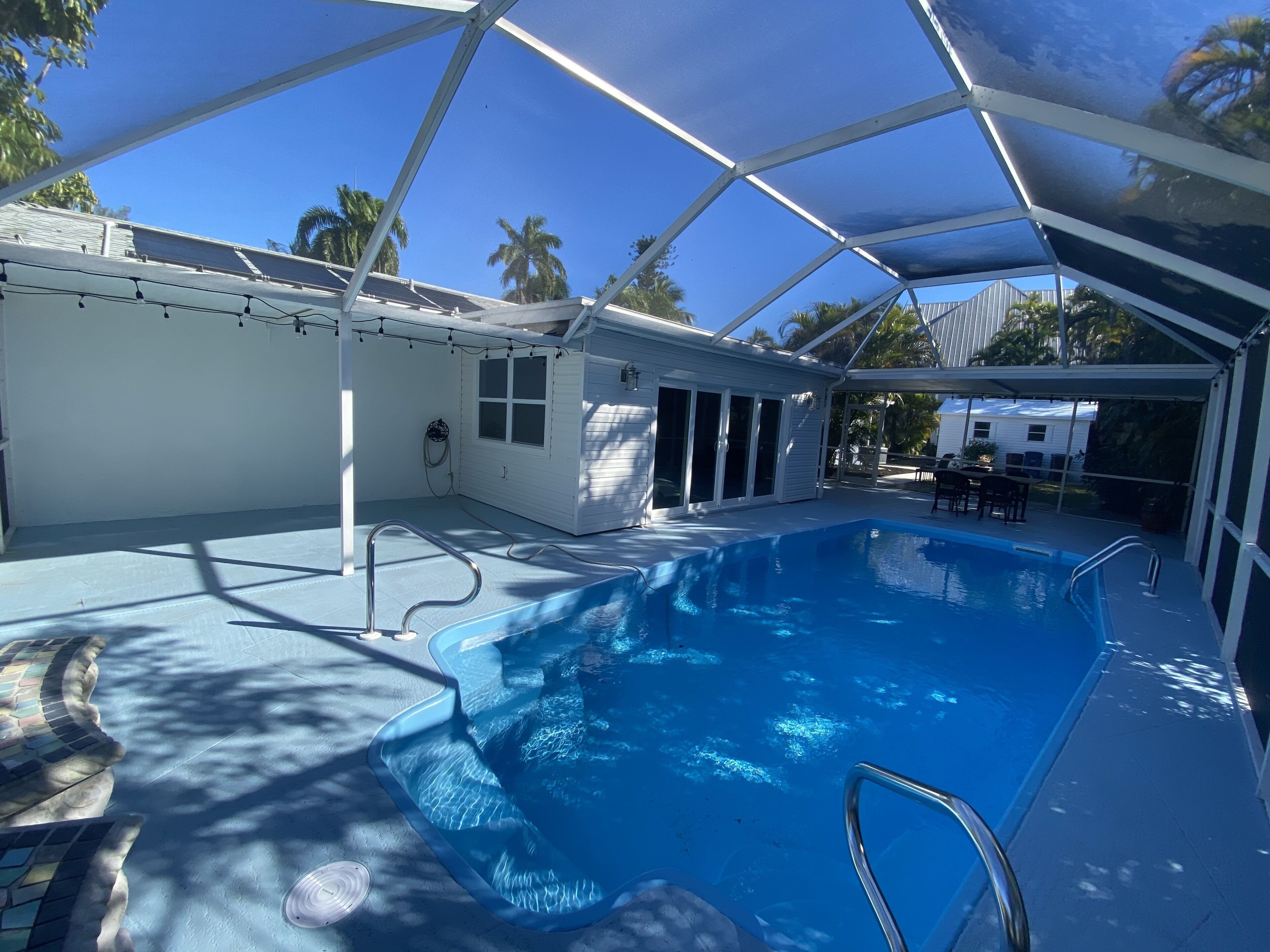 A covered swimming pool with a clear blue water feature, a white building with large windows, and palm trees in the background.