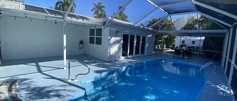 A covered swimming pool with a clear blue water feature, a white building with large windows, and palm trees in the background.