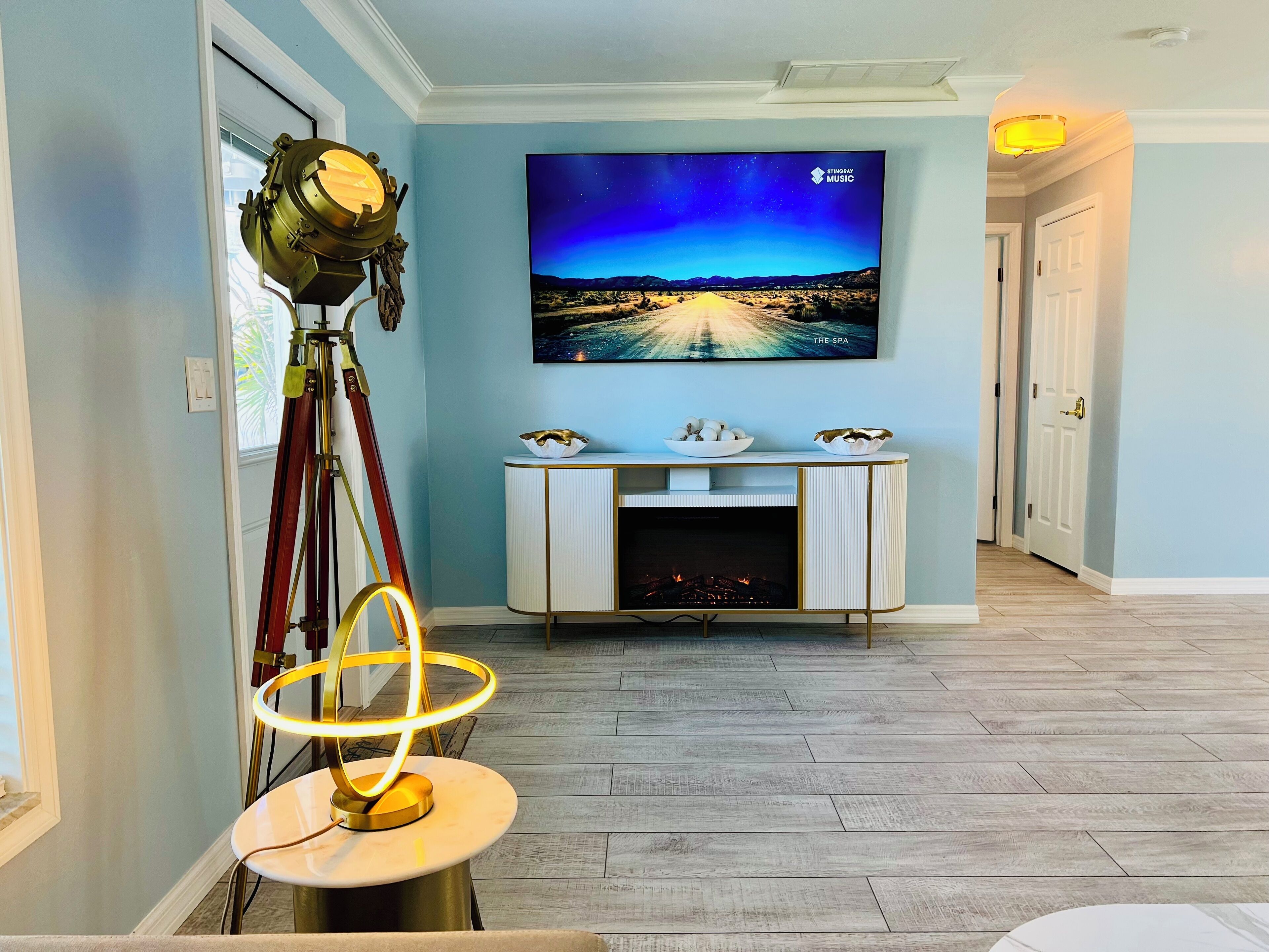 A living room with a mounted TV displaying 'The Spa' on the wall, a fireplace, a lamp, and a tripod with a light.